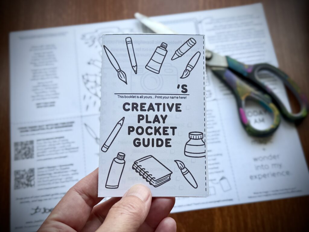 Close up of the cover of the JoeGirl Creative Play Pocket Guide, with full spread in the background but faded in behind, and a pair of paint-splattered scissors off to the side.